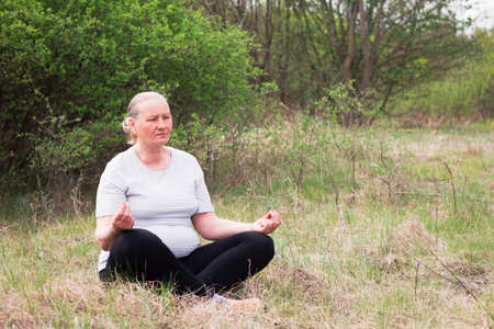 Portrait of a senior woman, wearing a white T-shirt and black pants, meditates in nature. Meditation, yoga and relaxation concept.の写真素材