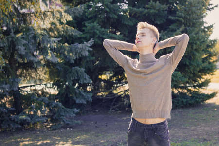 A young blond guy, dressed in a beige sweater and black jeans, stands with his eyes closed and holds his hands behind his head against the background of green fir trees.の写真素材