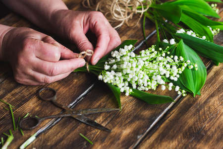 girl knits twine a bouquet of lilies of the valley on a wooden dark background, large stall. Florist makes a delicate bouquet of flowers.の写真素材