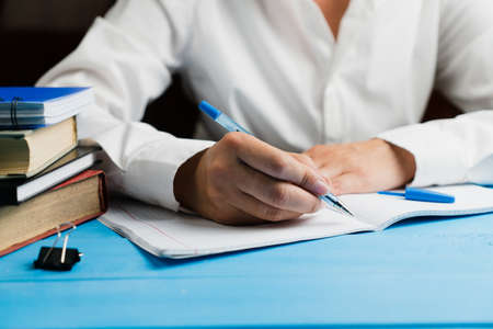 A schoolgirl in a white and clean shirt sits at a wooden table with an empty notebook and holds a blue pen with both hands. School concept.の写真素材