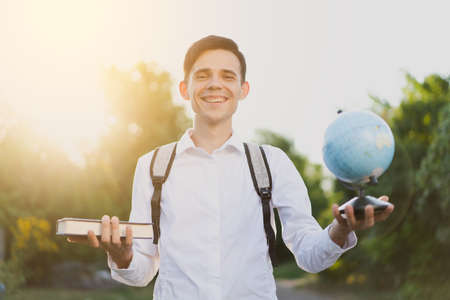 Smiling brunette guy with gray briefcase on his shoulders, dressed in  classic white shirt, stands and holds globe in one hand, and in the second book against the background of nature and sunset.の写真素材