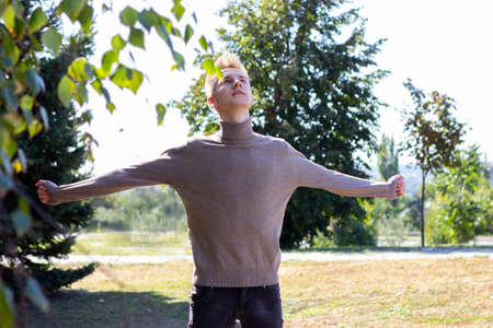 A young blond European guy, dressed in a bodysuit, stands with outstretched hands against the background of autumn nature. Photo concept.の写真素材