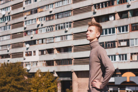 A young blond guy, dressed in a beige sweater and black jeans, stands half-sided with his eyes closed against the background of an ordinary multi-storey building.の写真素材