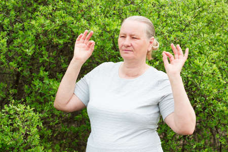 An athletic gray-haired woman, wearing a white T-shirt and black pants, meditates, holding her hands in the shape of approx. Aging, maturity, health concept.の写真素材