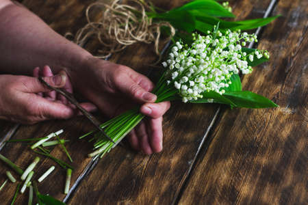 woman cuts the bottom of a bouquet of lilies of the valley. Making a bouquet of delicate white forest flowers with scissors and rope on a wooden backgroundの写真素材