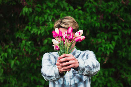 guy in a blue checkered shirt holds a bouquet of pink tulips holding them in front of him, close his face, outdoors.の写真素材