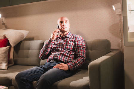 An adult bald man, dressed in a plaid shirt and jeans, is sitting on the couch and talking on a smartphone. Positive communication.の写真素材