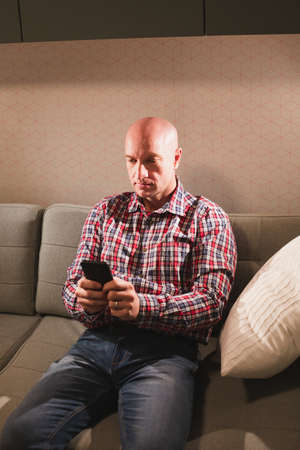 Portrait of a happy adult bald man, dressed in a plaid shirt and jeans, sitting on the couch with a phone.の写真素材