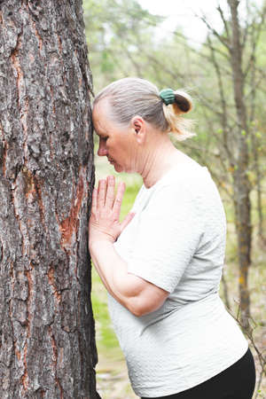 An adult woman, dressed in light-colored clothing, meditates by touching her head to a tree. She feels the sun and the atmosphere of a deciduous forest. Side view.の写真素材