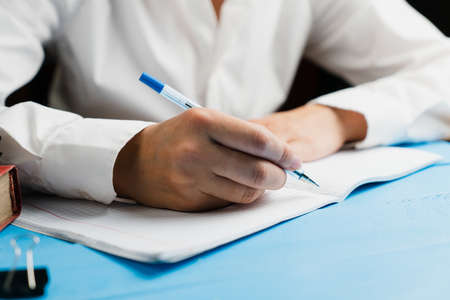 A schoolgirl in a white and clean shirt sits at a wooden table with an empty notebook and holds a blue pen with both hands. School concept.の写真素材