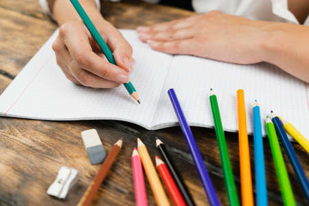 A schoolgirl, dressed in a white blouse, sits at a wooden desk and draws in a squared notebook with colored pencils. School concept.の写真素材