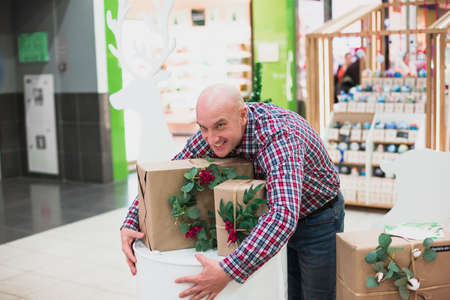 A good attractive bald man dressed in a plaid shirt and jeans looks at the presents and tries to hug them.の写真素材