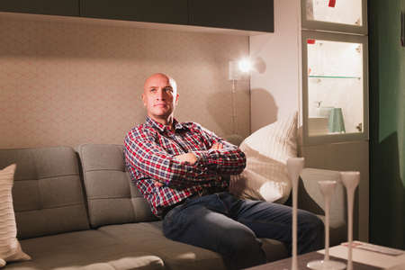 A carefree relaxed man dressed in a red plaid shirt and jeans is relaxing on a sofa in a cozy living room enjoying a lazy day off.の写真素材