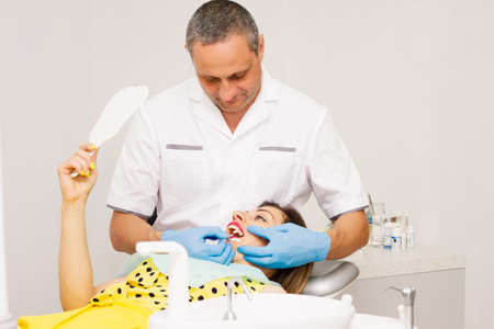 An adult male dentist, dressed in a clean white uniform and blue gloves, treats the teeth of a young woman in a yellow blouse. Clinic. Health concept.の写真素材