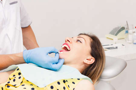 An attractive dark-haired woman, wearing a yellow polka-dot blouse, sits with her mouth open during a teeth whitening procedure. Health concept.の写真素材