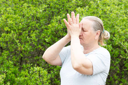 Happy mature woman, wearing a white t-shirt, meditates while standing and holding her hands near her head. Healthy life concept.の写真素材