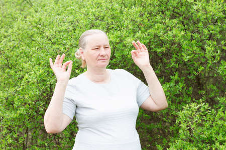 An athletic gray-haired woman, wearing a white T-shirt and black pants, meditates, holding her hands in the shape of approx. Aging, maturity, health concept.の写真素材