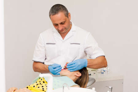 An adult male dentist, dressed in a clean white uniform and blue gloves, treats the teeth of a young woman in a yellow blouse. Clinic. Health concept.の写真素材