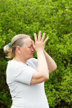Happy mature woman, wearing a white t-shirt, meditates while standing and holding her hands near her head. Healthy life concept.の写真素材