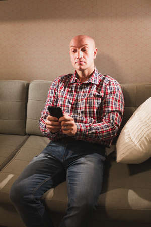 Portrait of a happy adult bald man, dressed in a plaid shirt and jeans, sitting on the couch with a...