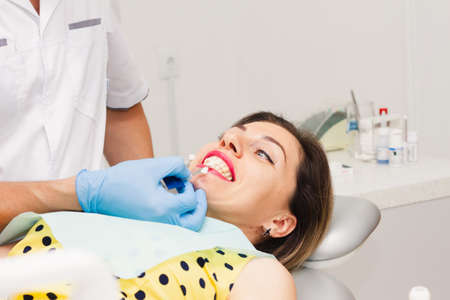 An attractive dark-haired woman, wearing a yellow polka-dot blouse, sits with her mouth open during a teeth whitening procedure. Health concept.の写真素材