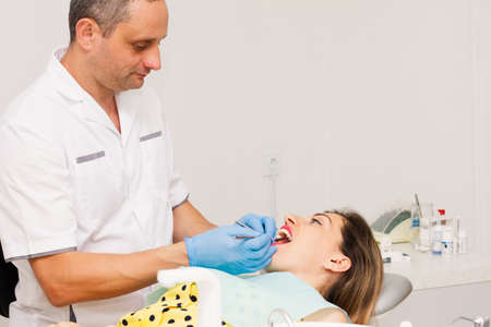 An adult male dentist, dressed in a clean white uniform and blue gloves, treats the teeth of a young woman in a yellow blouse. Clinic. Health concept.の写真素材