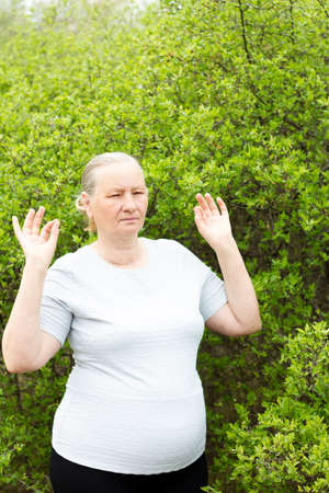 An athletic gray-haired woman, wearing a white T-shirt and black pants, meditates, holding her hands in the shape of approx. Aging, maturity, health concept.の写真素材
