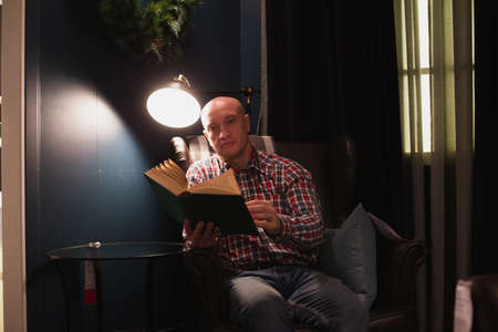 An adult man learns by reading a book in focus at night by the light of a lamp. A dark room.の写真素材