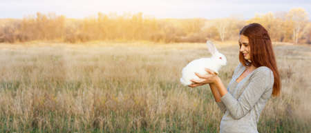 young girl with a red hair and a gray jacket is holding  small white fluffy rabbit with red eyes in front of her, smiling sweetly at him. With space for your text. On a dry steppe area. Easter conceptの写真素材