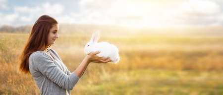 young girl with a red hair and a gray jacket is holding  small white fluffy rabbit with red eyes in front of her, smiling sweetly at him. With space for your text. On a dry steppe area. Easter conceptの写真素材