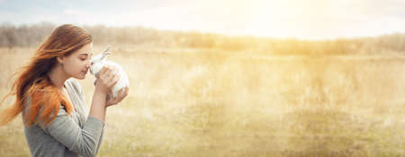 young girl with a red hair and a gray jacket is holding  small white fluffy rabbit with red eyes in front of her, smiling sweetly at him. With space for your text. On a dry steppe area. Easter conceptの写真素材