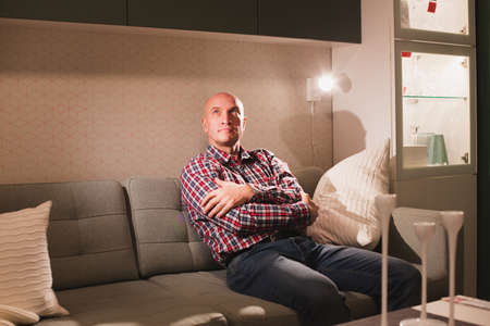 A carefree relaxed man dressed in a red plaid shirt and jeans is relaxing on a sofa in a cozy living room enjoying a lazy day off.の写真素材