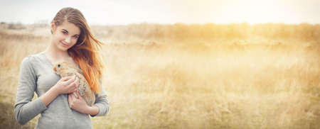 a young woman with red hair and in a gray jacket of Caucasian ethnicity holds a light red rabbit in her arms and looks into the camera. Place for your text. Banner photo. Spring holidays conceptの写真素材