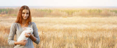 a young woman with red hair and a gray jacket of Caucasian ethnicity holds a white rabbit in her arms and looks into the camera. Place for your text. Banner photo. Spring holidays conceptの写真素材
