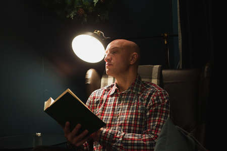 An adult man learns by reading a book in focus at night by the light of a lamp. A dark room.の写真素材