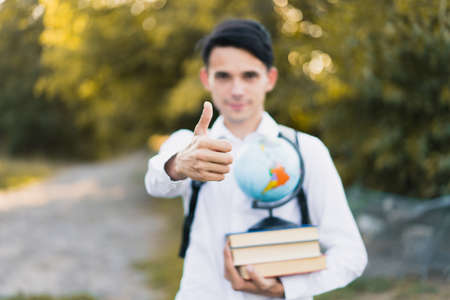 A young handsome guy in a white classic shirt holds books with a globe in his hand and shows a thumb up on his outstretched hand. Selective focus. On open air.の写真素材