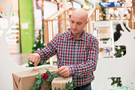 A good attractive bald man dressed in a plaid shirt and jeans looks at the presents and tries to hug them.の写真素材