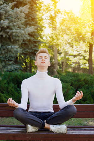 A young blond guy, dressed in a white sweater and black jeans, sits in a lotus position on a bench against the backdrop of a city park. Health concept.の写真素材