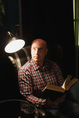 A man, dressed in a plaid shirt and jeans, sits in an armchair and reads a book at dusk. Artificial lighting. Life concept.の写真素材