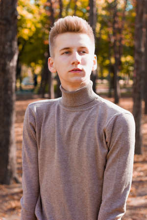 A young blond guy, dressed in a beige sweater and black jeans, posing against the backdrop of an autumn city park. Photo concept.の写真素材