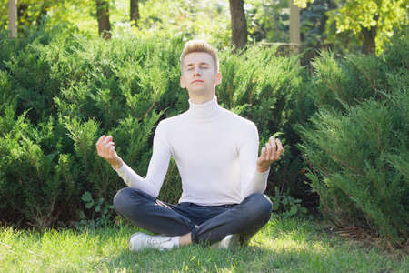 A young blond guy, dressed in a white sweater and black jeans, sits in a lotus position with closed eyes on the background of nature. Health concept.の写真素材