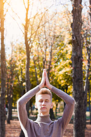 A young blond guy, dressed in a beige sweater, stands with his eyes closed, stretching his arms up and folding them against the background of nature.の写真素材