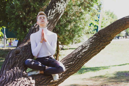 A young blond guy, dressed in a white sweater and black jeans, sits on a tree with his legs and arms together in the park. Photo concept.の写真素材
