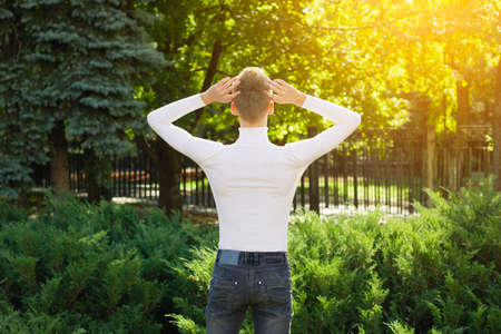 A young blond guy, dressed in a white sweater and black jeans, stands with his back to the camera, holding his head in his hands against the backdrop of a city park. Photo concept.の写真素材