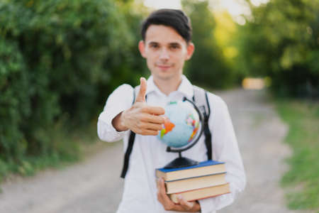 A young handsome guy in a white classic shirt holds books with a globe in his hand and shows a thumb up on his outstretched hand against the background of nature. Selective focus. On open air.の写真素材
