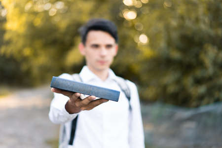 A young handsome guy in a white dress shirt holds a book in his hand and holds it out. Selective focus. On open air.の写真素材