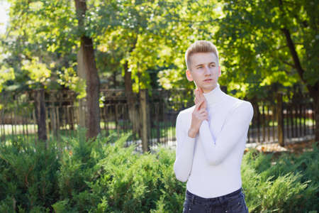 A young blond guy, dressed in a white sweater and black jeans, posing against the backdrop of a city park. Photo concept.の写真素材