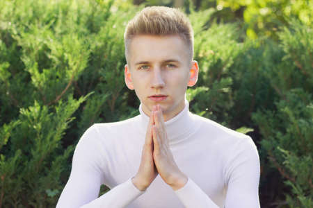 A young blond guy, dressed in a white jacket, sits and holds his hands clasped against the background of the park.の写真素材