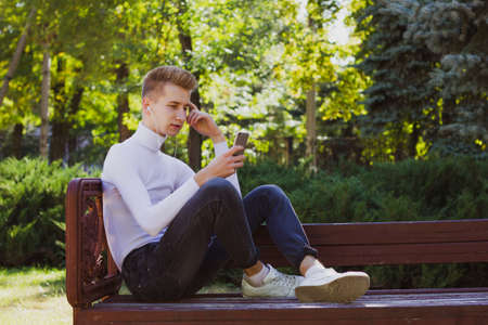 A young blond guy, dressed in a white jacket and black jeans, sits on a bench with headphones and looks at the phone against the background of the park. Life concept.の写真素材