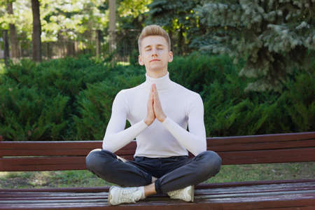 A young blond guy, dressed in a white sweater and black jeans, sits with his legs and arms together against the backdrop of a city park. Photo concept.の写真素材